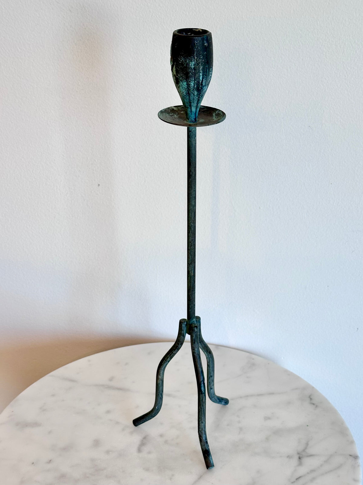 A dark green patina metal candlestick holder with a three-legged stand and a single cup, displayed on a round white marble table against a white wall., photo 5