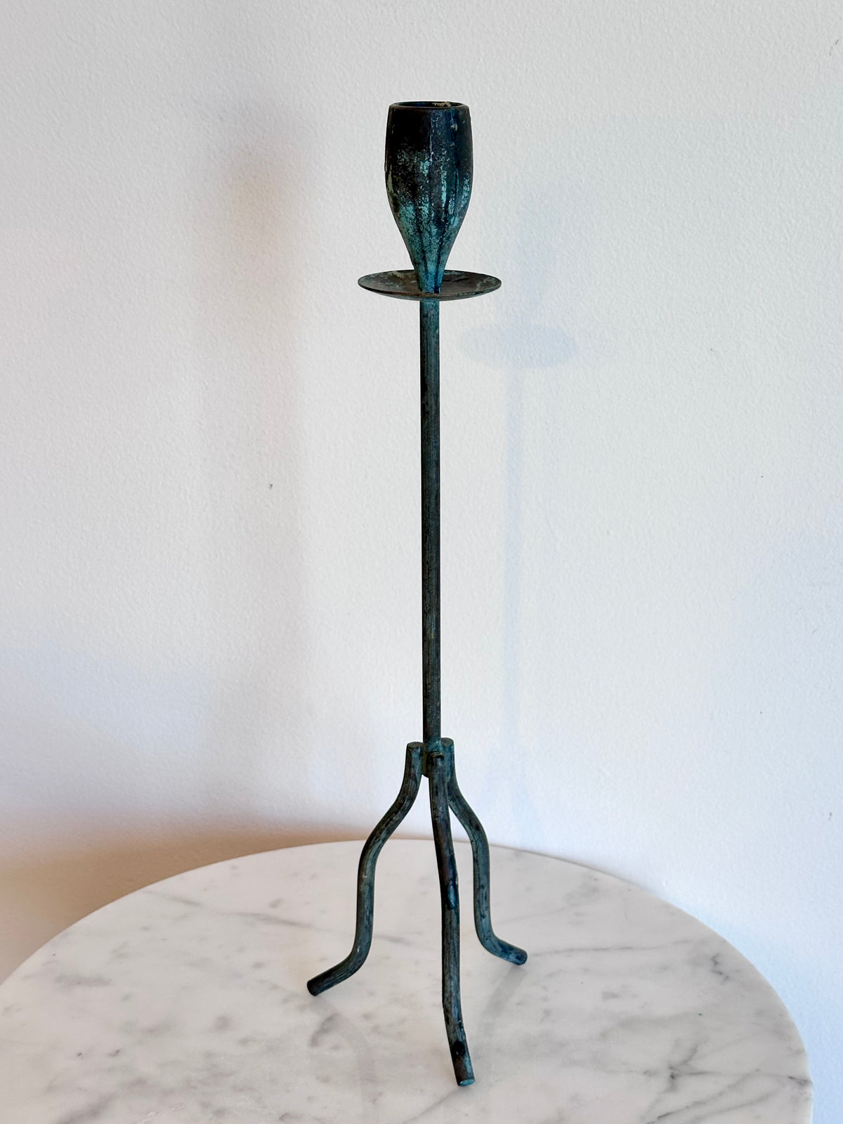 A dark green patina metal candlestick holder with a three-legged stand base and a single stem holding a cup-shaped candle holder, displayed on a white marble round table against a white wall., photo 6