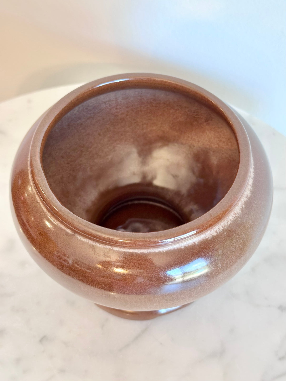 A round, glossy ceramic pot or planter in a warm brown glaze, photographed from a top-down angle on a light marble surface., photo 5