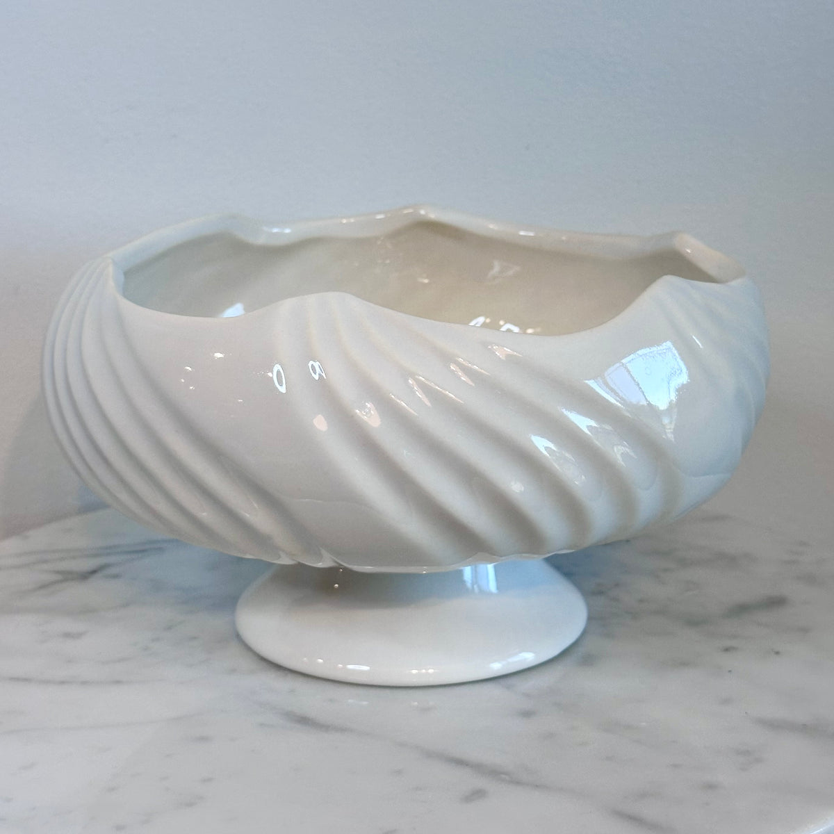 A glossy white ceramic pedestal bowl with a fluted, ribbed exterior and wavy rim, displayed on a white marble surface against a light gray background.