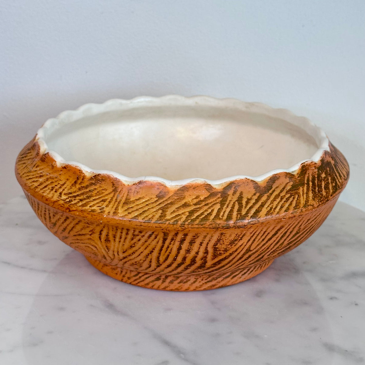 A round ceramic bowl with a textured, ribbed exterior in a warm orange-brown glaze and a scalloped, glossy off-white interior, resting on a white marble surface against a light gray background.