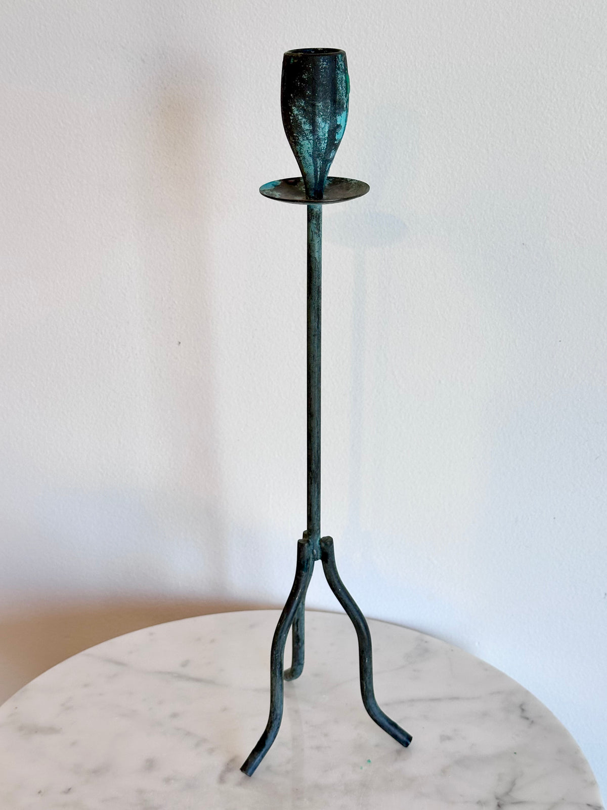 A tall, slender metal candlestick holder with a three-legged tripod base and a textured, dark teal-green finish, standing on a round white marble surface against a plain white wall.