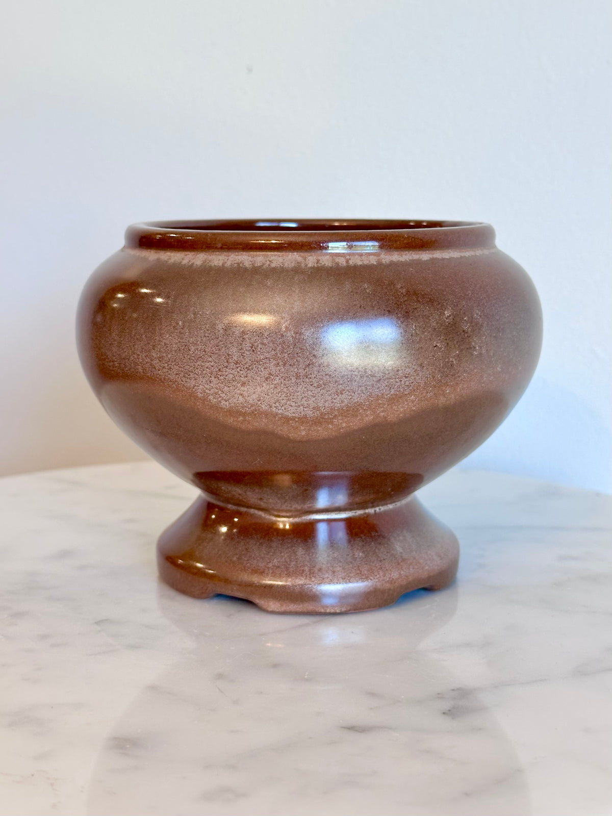 A small round ceramic planter pot with a  brown and bronze glaze, sitting on a white marble surface against a plain white wall.