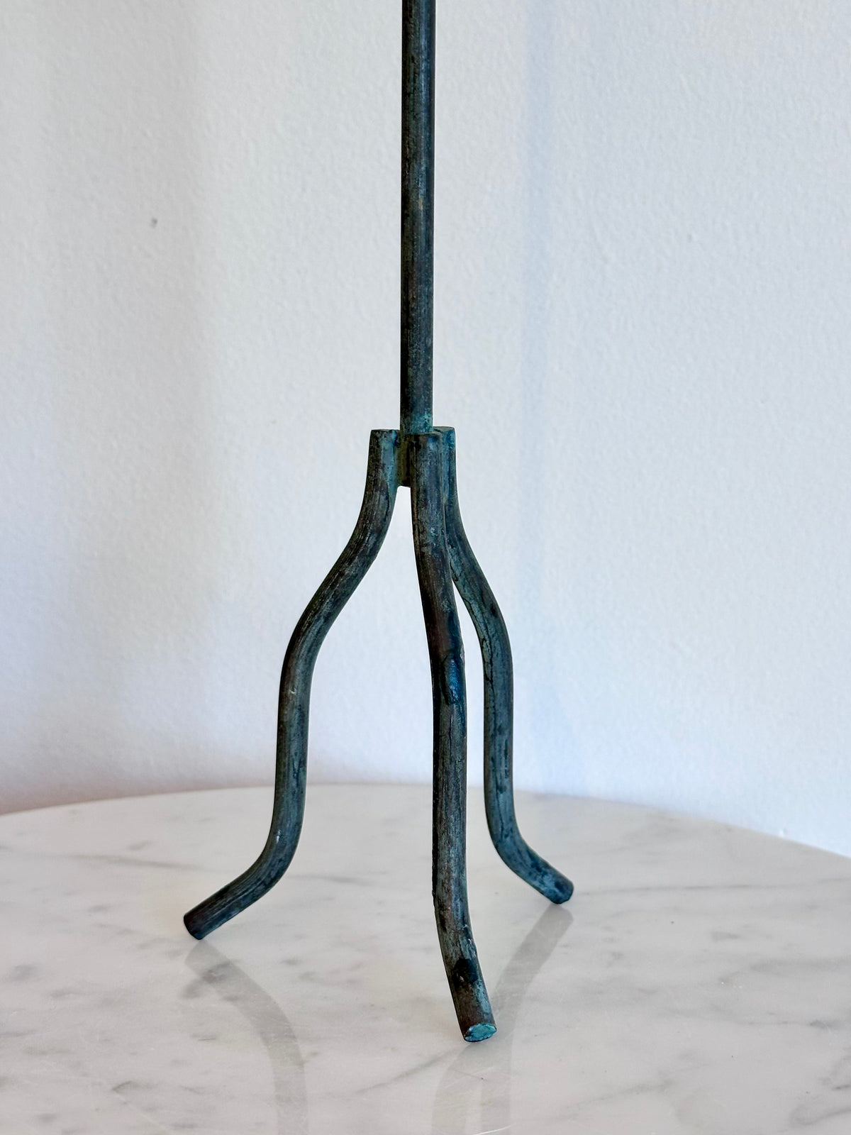 A dark metal stand with a three-legged tripod base and a vertical stem, featuring a distressed teal and black patina finish, displayed on a white marble surface against a white wall., photo 3