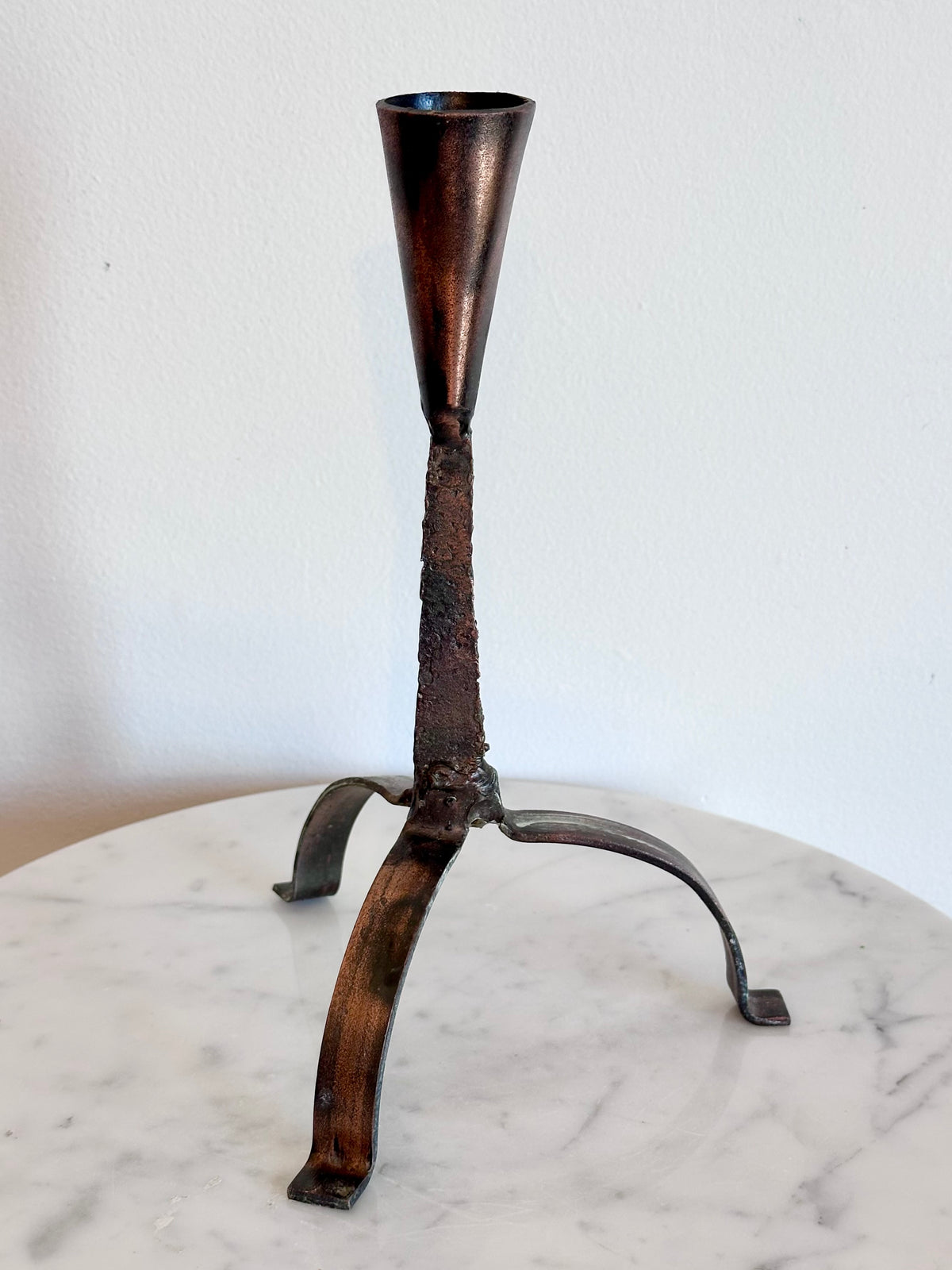 A rustic metal candle holder with a conical top and three curved legs, shown standing on a white marble surface against a plain white wall.