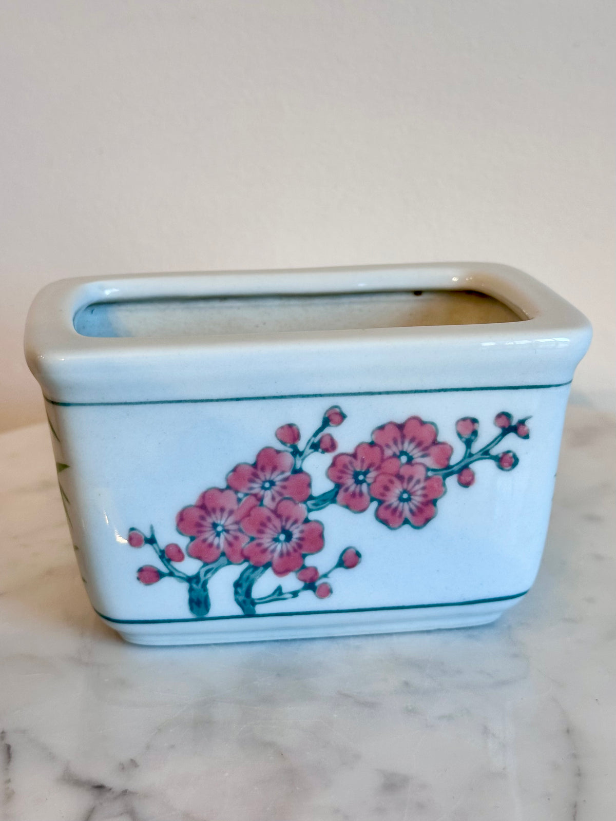A rectangular ceramic tissue box cover with a white glaze, decorated with a pink cherry blossom branch motif and a thin teal border, shown on a marble surface against a plain beige background., photo 5