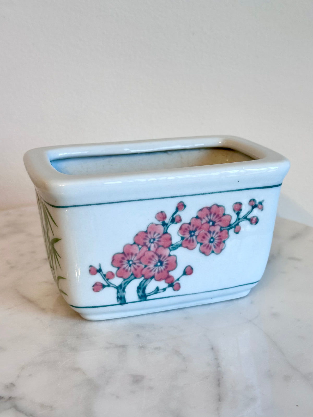 A rectangular white ceramic planter with a glossy finish, decorated with pink cherry blossoms and green leaves, sitting on a white marble surface against a plain beige wall.