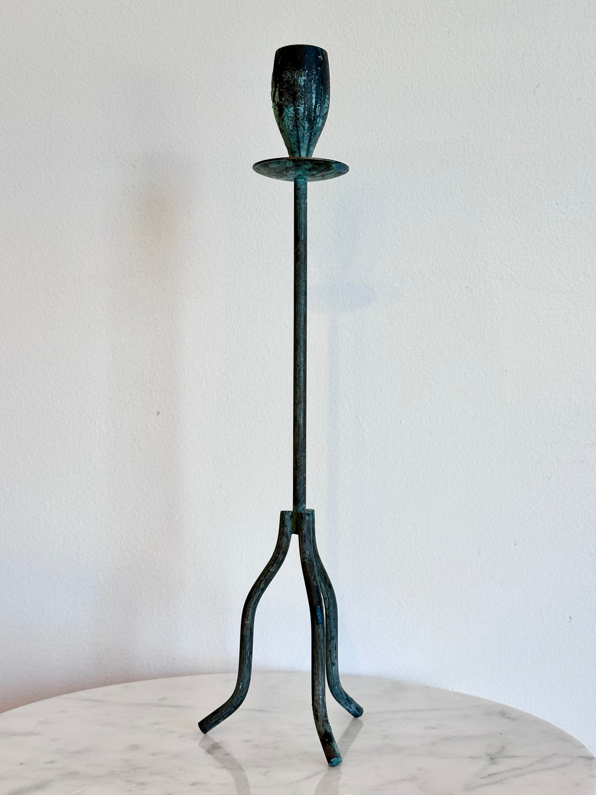 A tall, slender metal candle holder with a three-legged stand and a single cup-shaped holder at the top, finished in a dark green and black oxidized patina, displayed on a round white marble surface against a white wall., photo 2