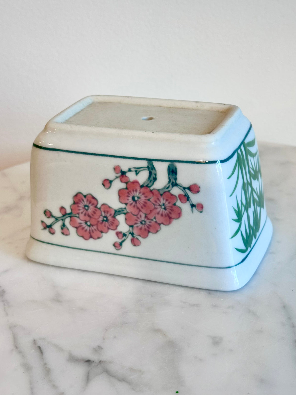 A small square white ceramic planter or pot with a painted floral design featuring pink blossoms and green leaves, shown on a white marble surface against a plain light background., photo 6