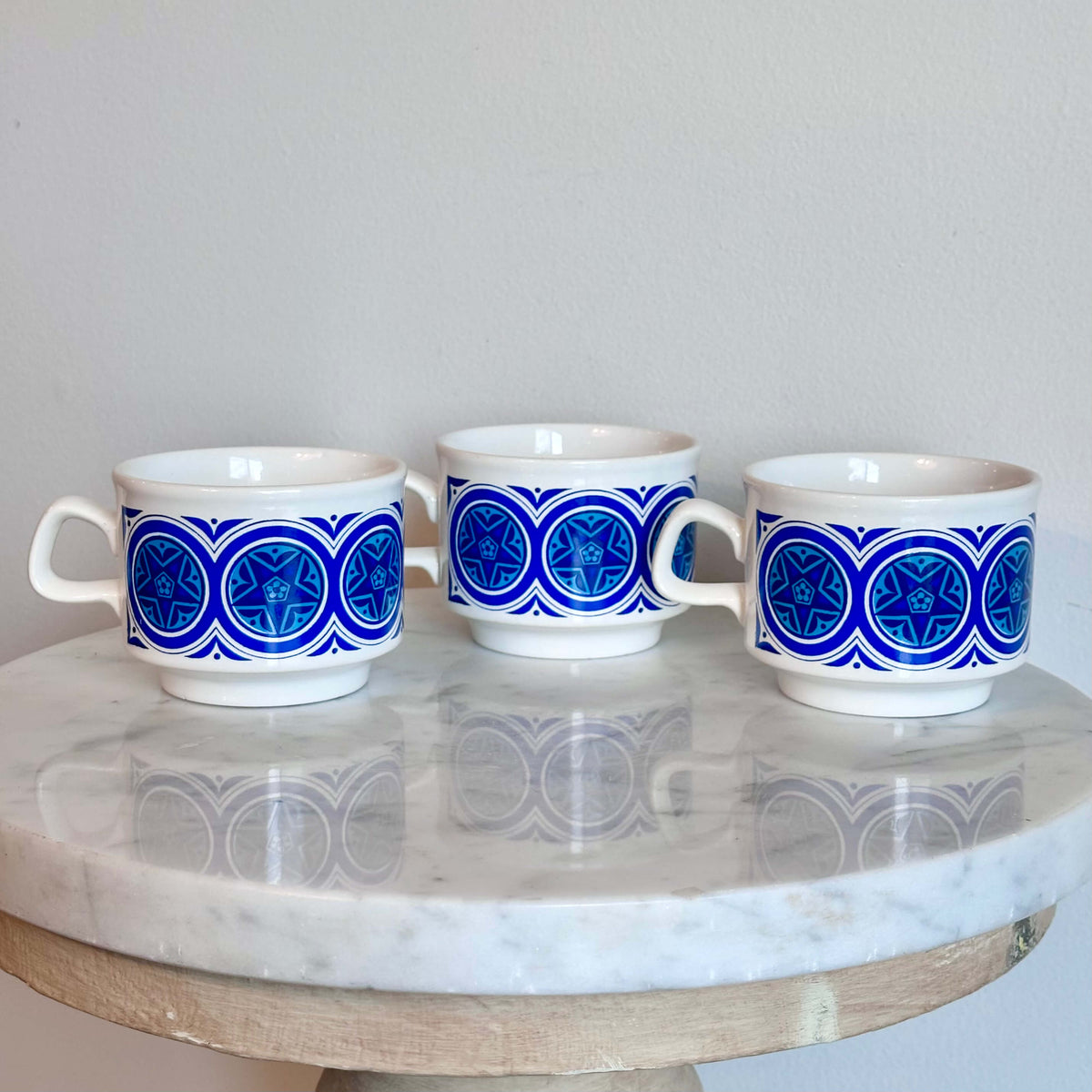 Three white ceramic cups with blue geometric patterns, arranged on a round white marble table.