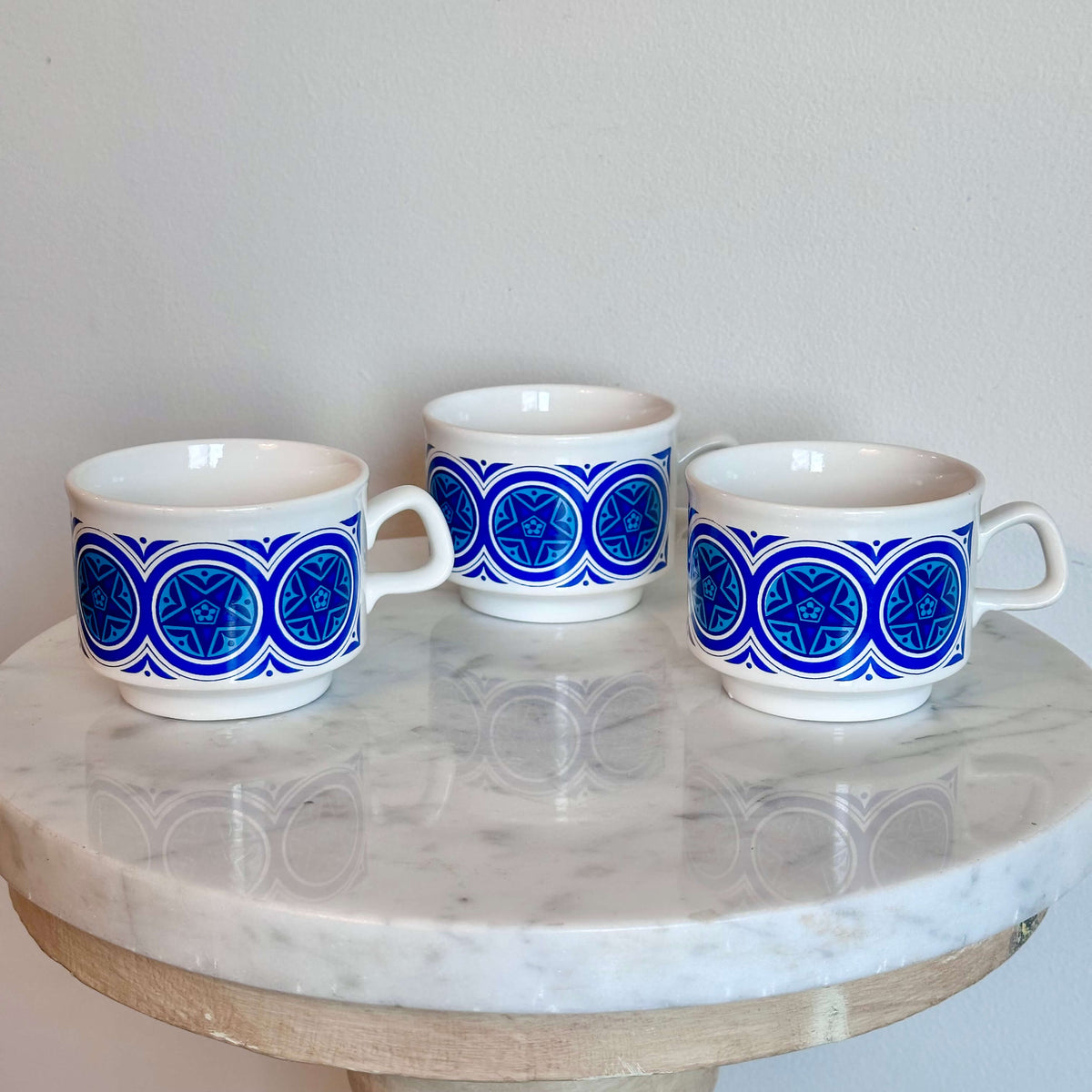 Three white ceramic cups with blue geometric medallion patterns, arranged on a round white marble table.