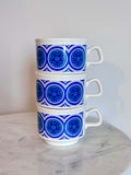 Three stacked white ceramic mugs with a blue geometric circular pattern, set on a white marble surface against a plain light gray wall.