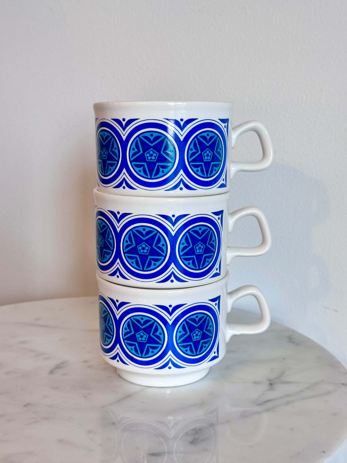 Three stacked white ceramic mugs with a blue geometric circular pattern, set on a white marble surface against a plain light gray wall.
