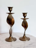 Two vintage brass spiral candlesticks of different heights displayed on a white marble surface against a white wall.