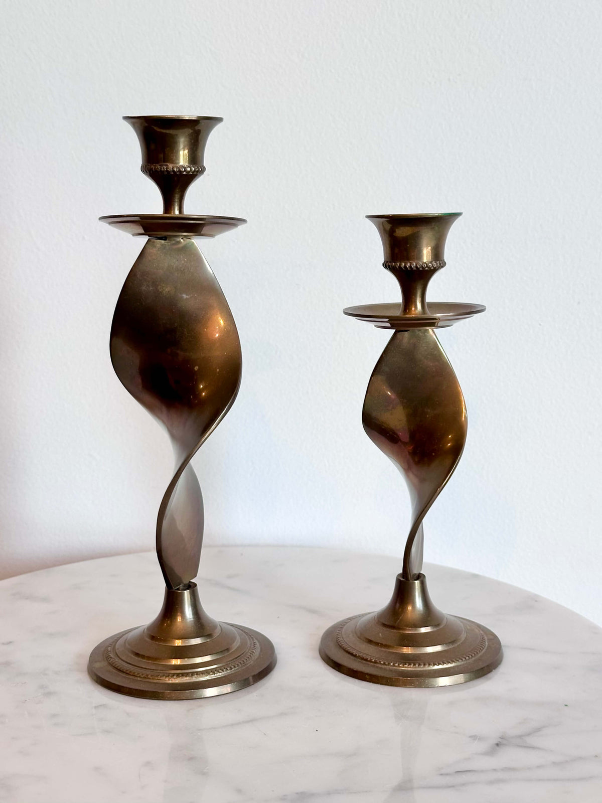 Two vintage brass spiral candlesticks of different heights displayed on a white marble surface against a white wall.