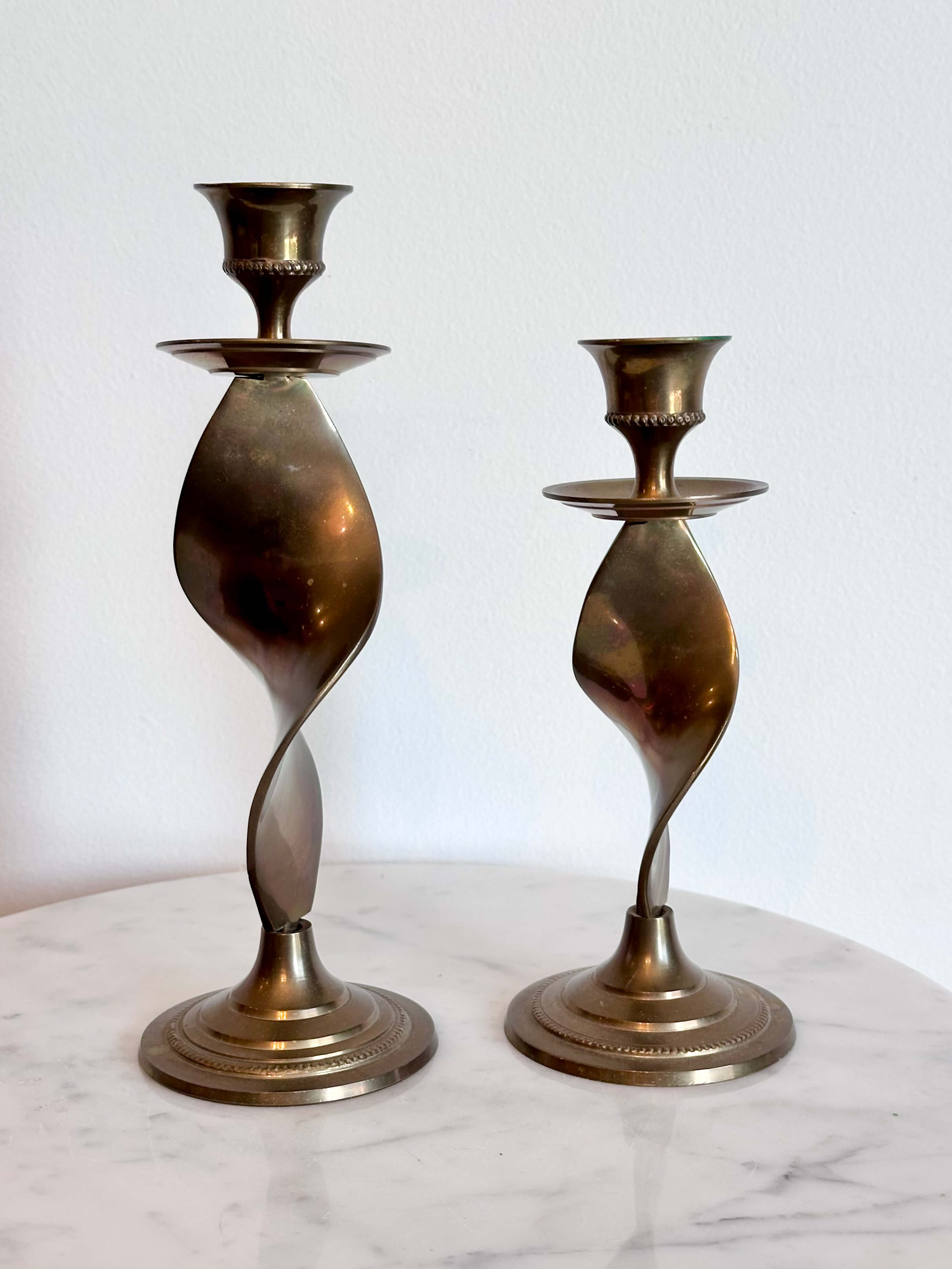 Two vintage brass spiral candlesticks of different heights displayed on a white marble surface against a white wall.