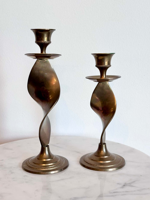 Two vintage brass spiral candlesticks of different heights displayed on a white marble surface against a white wall.