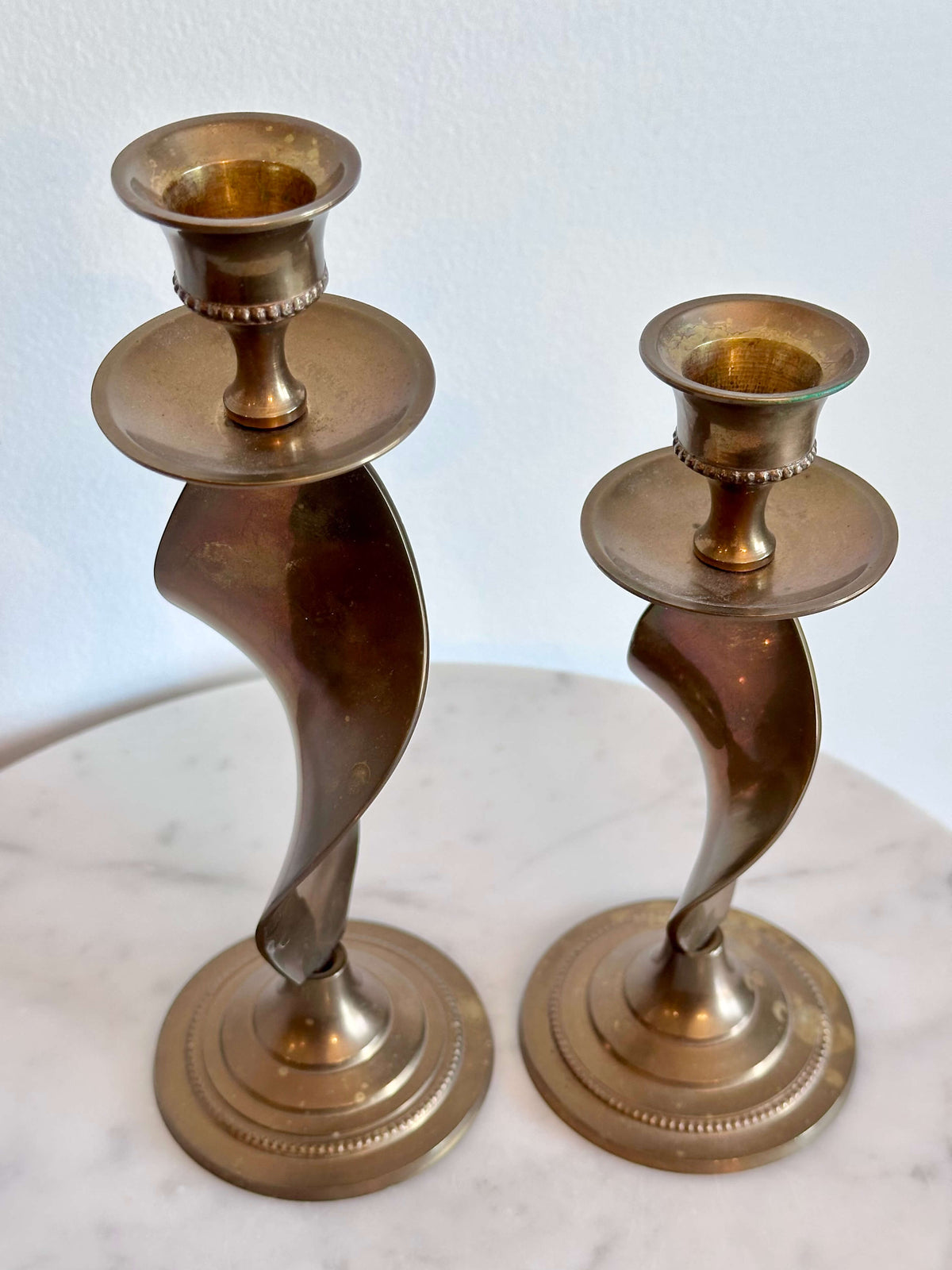 A pair of vintage brass spiral candlesticks with circular bases, displayed on a white marble surface against a white wall.