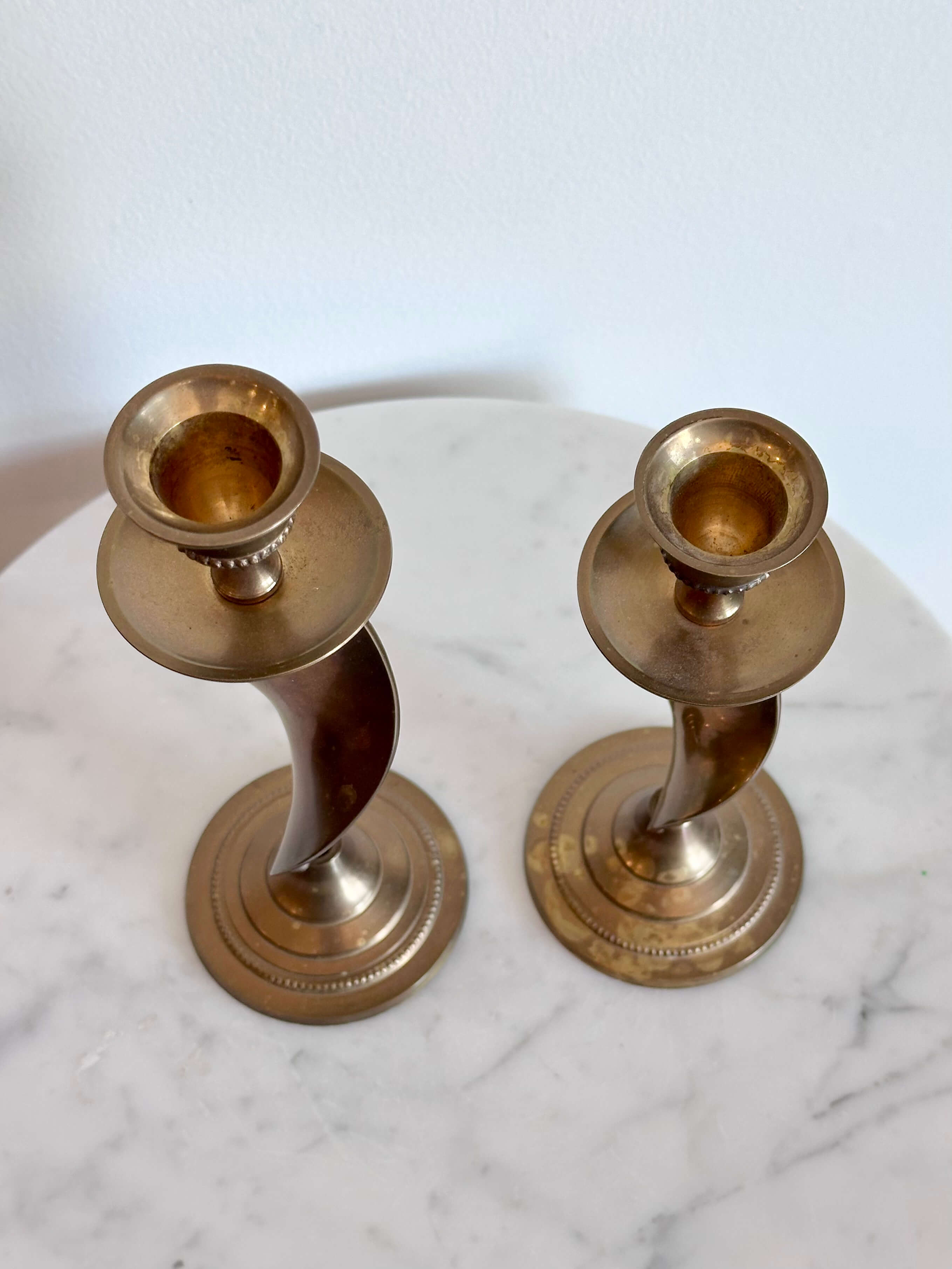 A pair of vintage brass spiral candlesticks with circular bases, displayed on a white marble surface against a white wall.
