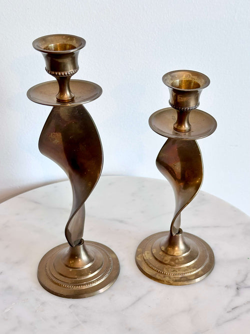 A pair of vintage brass spiral candlesticks with circular bases, displayed on a white marble surface against a white wall.