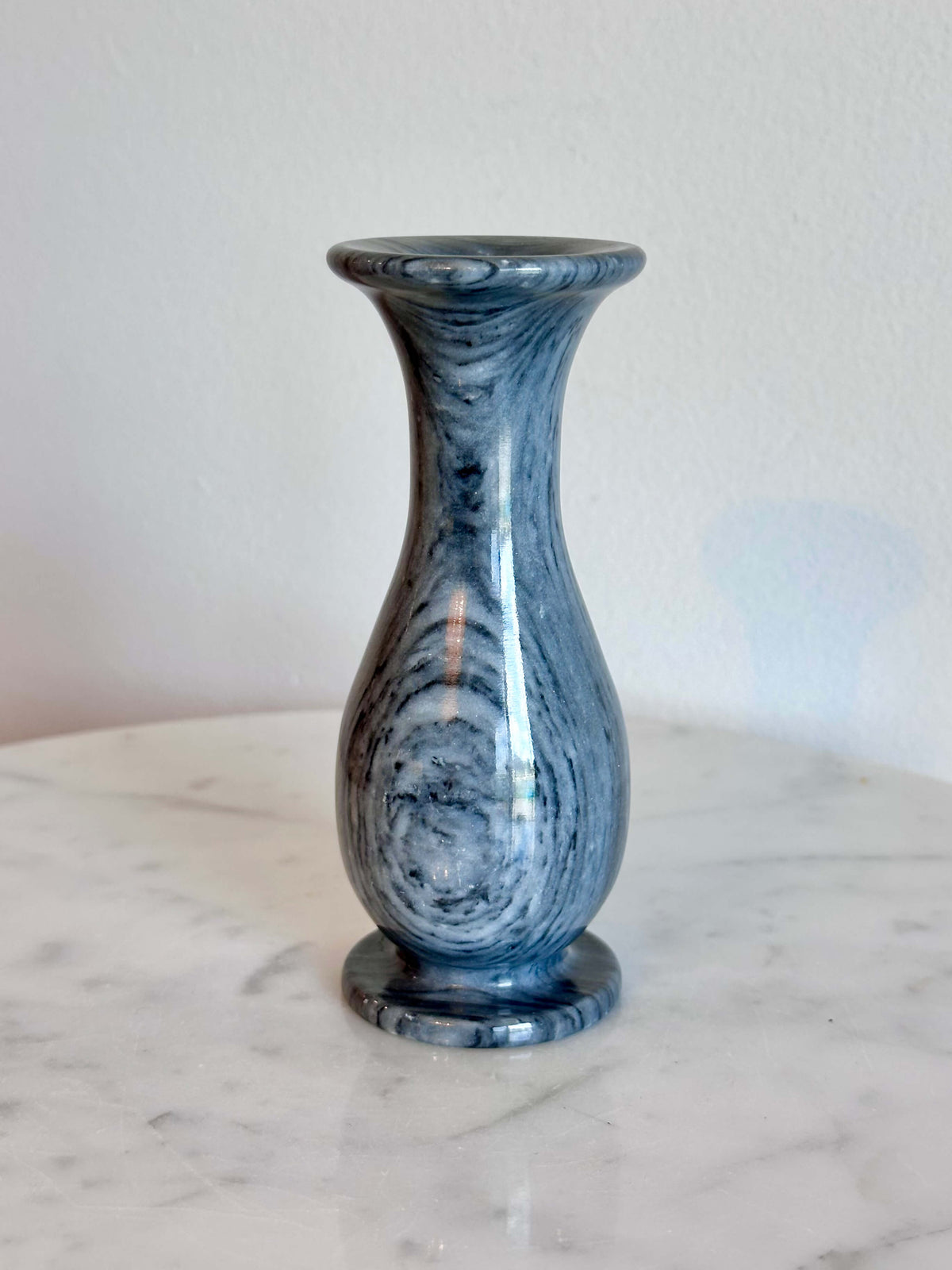 A small gray and white marbled stone vase with a flared rim and circular base, standing on a white marble surface against a plain off-white wall.