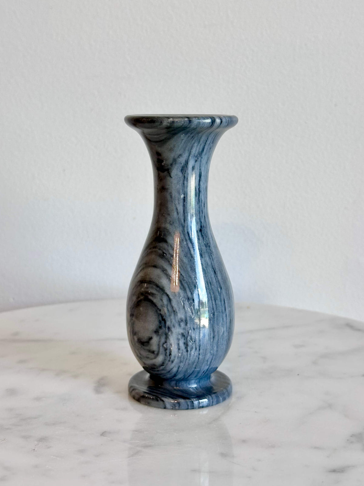 A small gray and white marbled stone vase with a flared rim and circular base, standing on a white marble surface against a plain off-white wall.