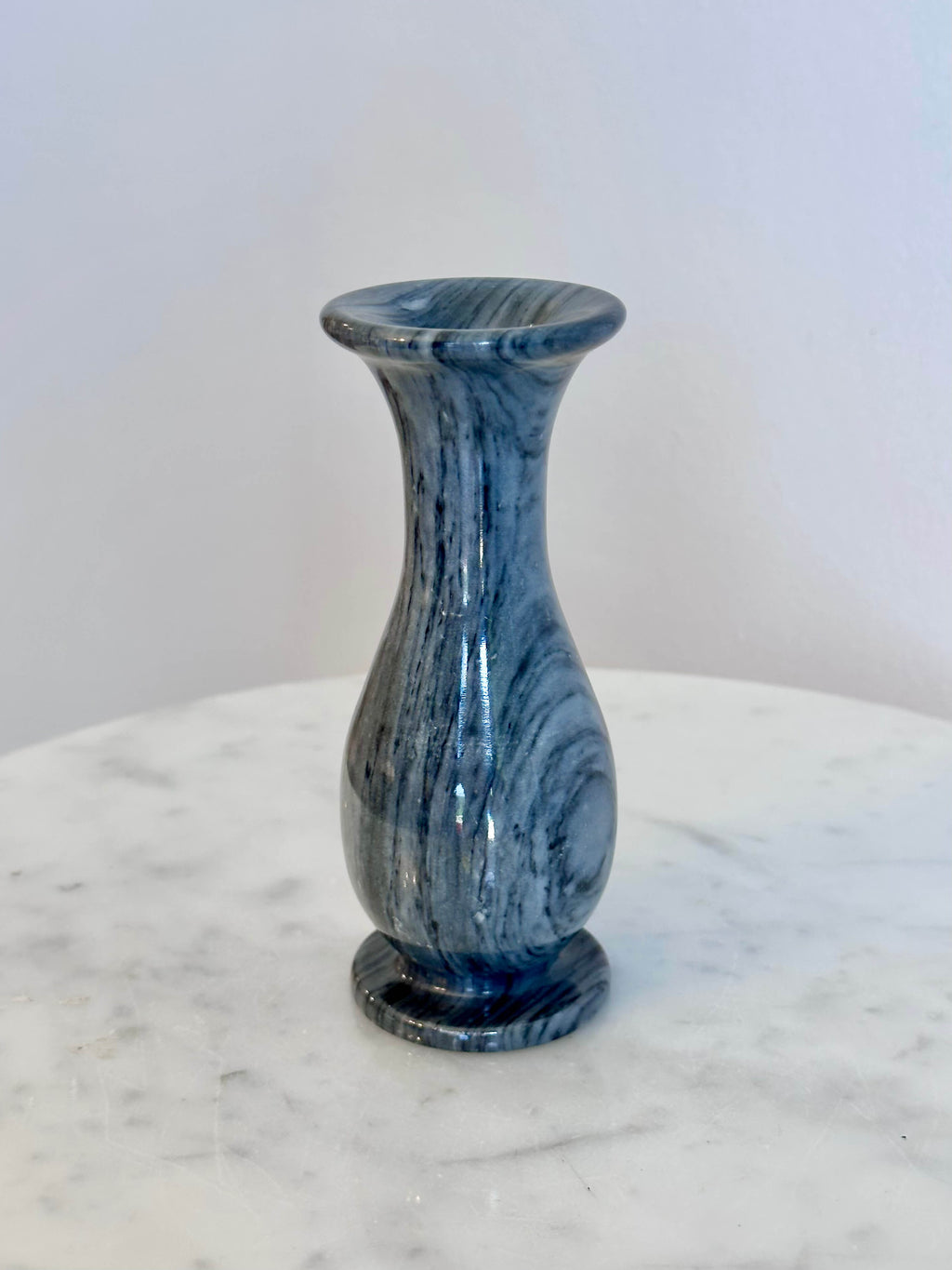 A small gray and white marbled stone vase with a flared rim and circular base, standing on a white marble surface against a plain off-white wall.