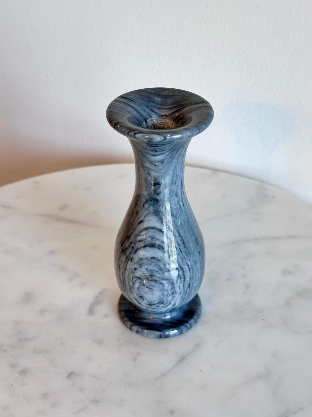 A small gray and white marbled stone vase with a flared rim and circular base, standing on a white marble surface against a plain off-white wall.