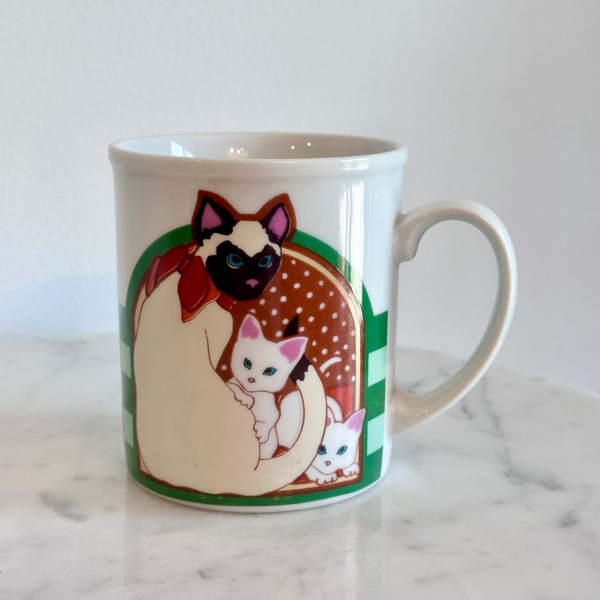 A white ceramic coffee mug featuring a colorful illustration of a Siamese cat holding two white kittens, set on a white marble surface against a white wall.
