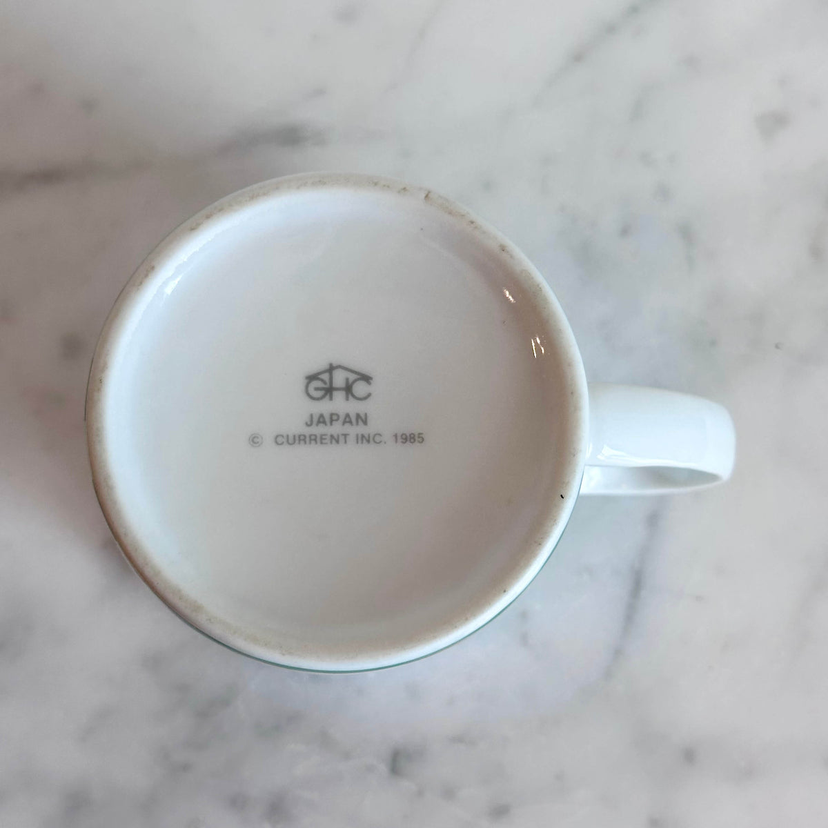 A white ceramic coffee mug featuring a colorful illustration of a Siamese cat holding two white kittens, set on a white marble surface against a white wall.