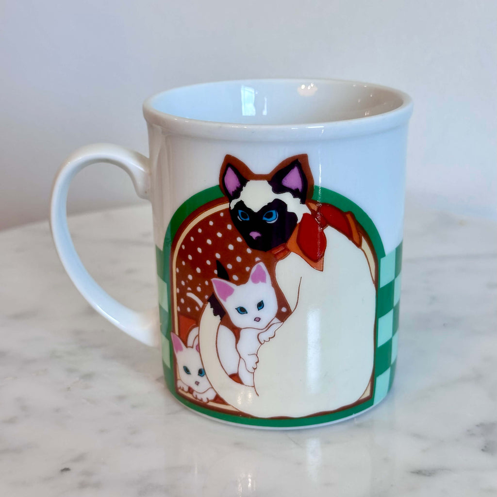 A white ceramic coffee mug featuring a colorful illustration of a Siamese cat holding two white kittens, set on a white marble surface against a white wall.