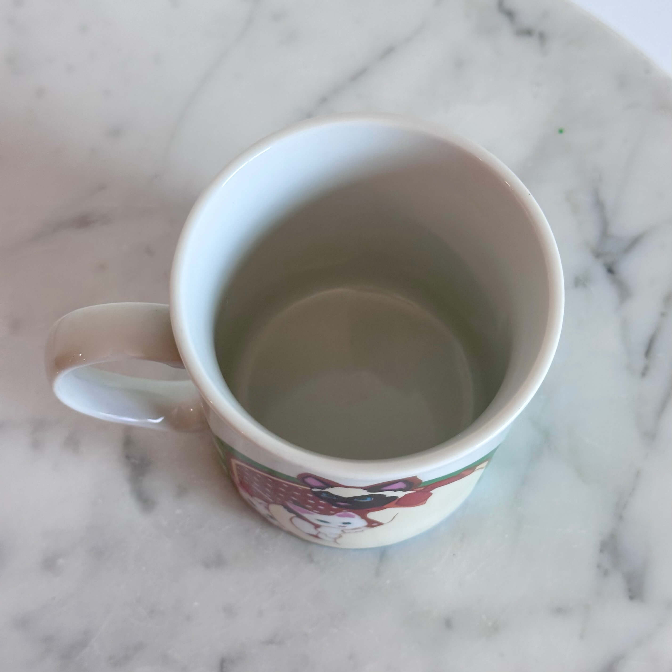 A white ceramic coffee mug featuring a colorful illustration of a Siamese cat holding two white kittens, set on a white marble surface against a white wall.