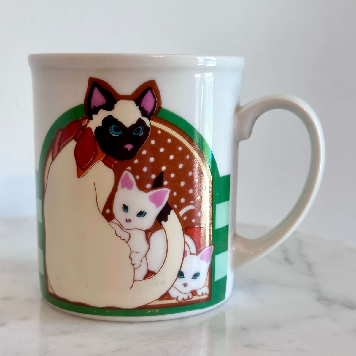 A white ceramic coffee mug featuring a colorful illustration of a Siamese cat holding two white kittens, set on a white marble surface against a white wall.