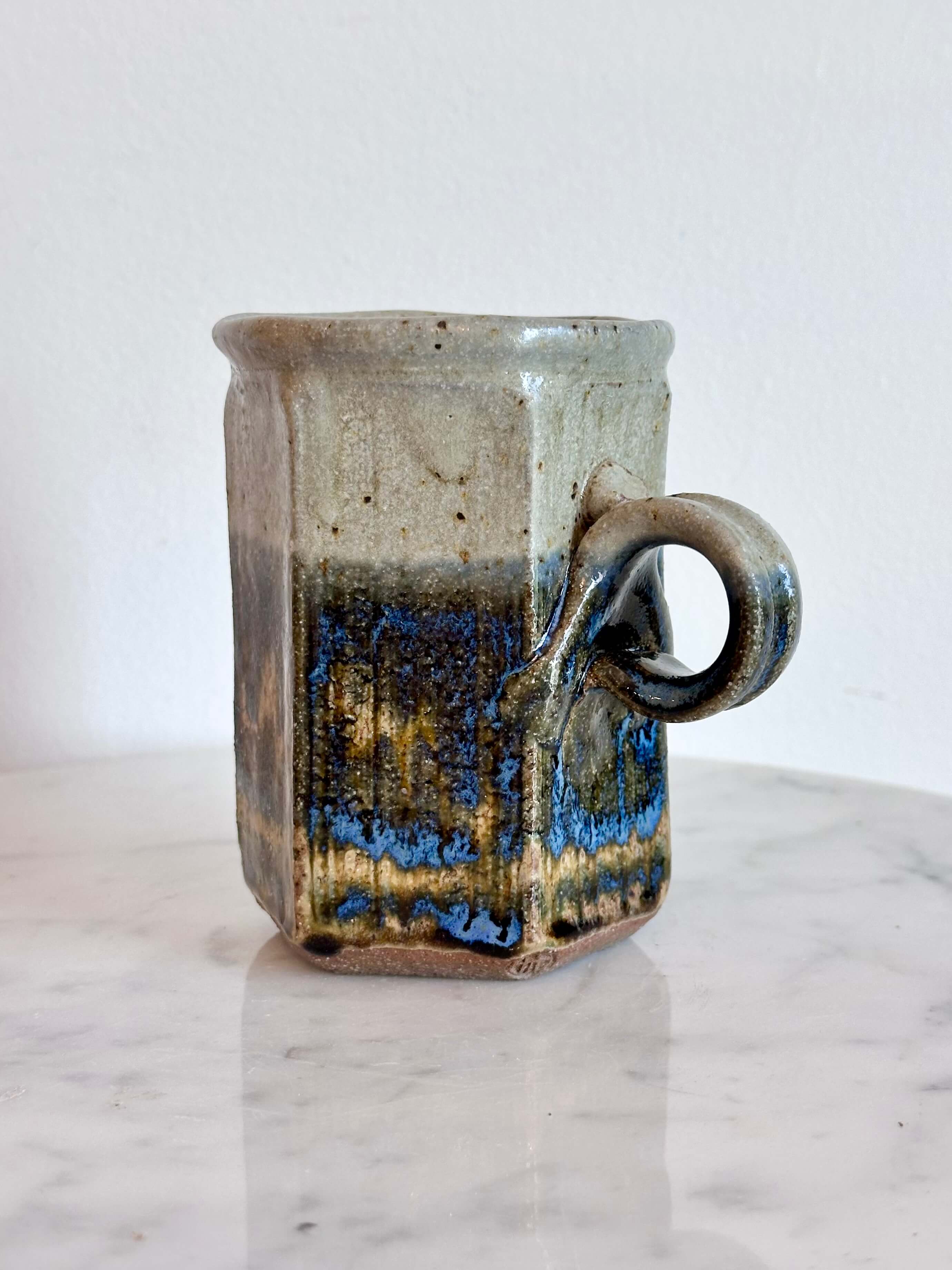 A handmade ceramic mug with a hexagonal body and a loop handle, featuring a layered glaze in gray, blue, and brown.