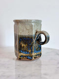 A handmade ceramic mug with a hexagonal body and a loop handle, featuring a layered glaze in gray, blue, and brown.