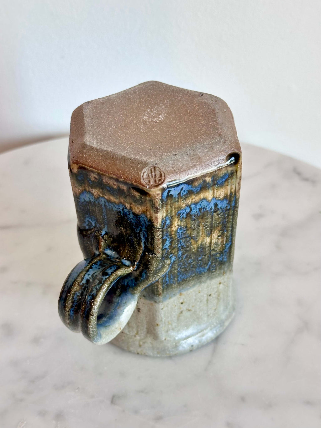 Bottom view of a handmade ceramic mug with a hexagonal body and a loop handle, featuring a layered glaze in gray, blue, and brown.