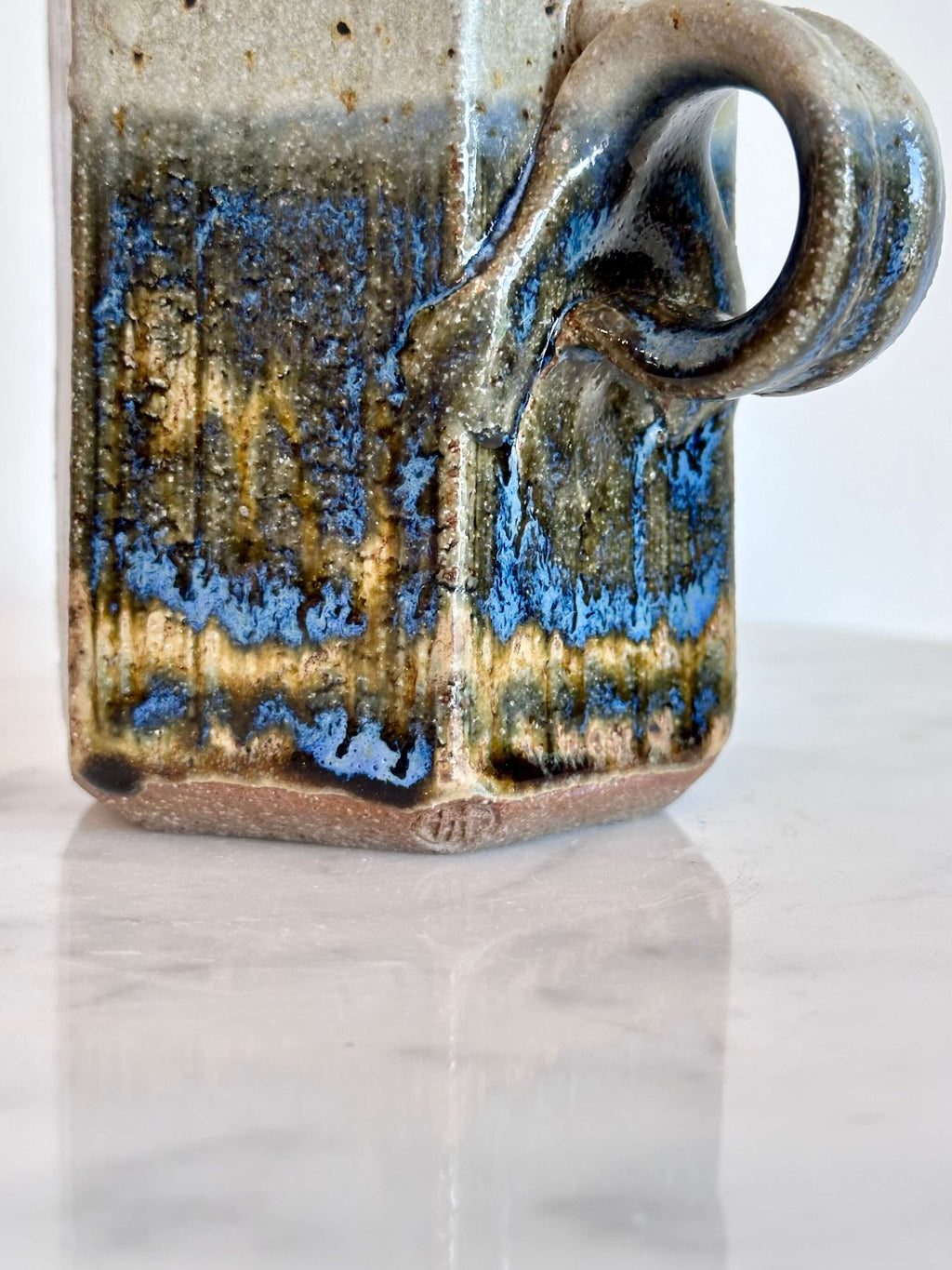 Close up of artist's stamp and handle of handmade ceramic mug with a hexagonal body and a loop handle, featuring a layered glaze in gray, blue, and brown.