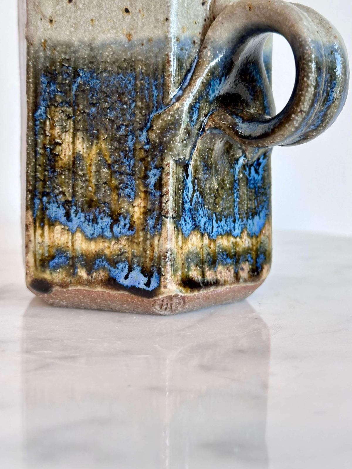 Close up of artist's stamp and handle of handmade ceramic mug with a hexagonal body and a loop handle, featuring a layered glaze in gray, blue, and brown.