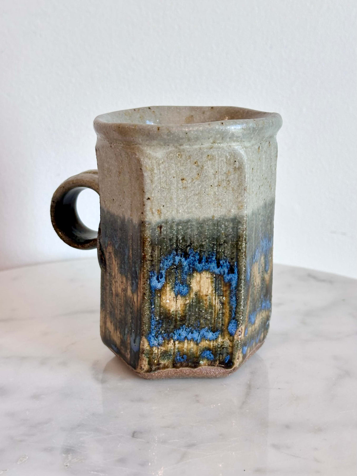 A handmade ceramic mug with a hexagonal body and a loop handle, featuring a layered glaze in gray, blue, and brown.