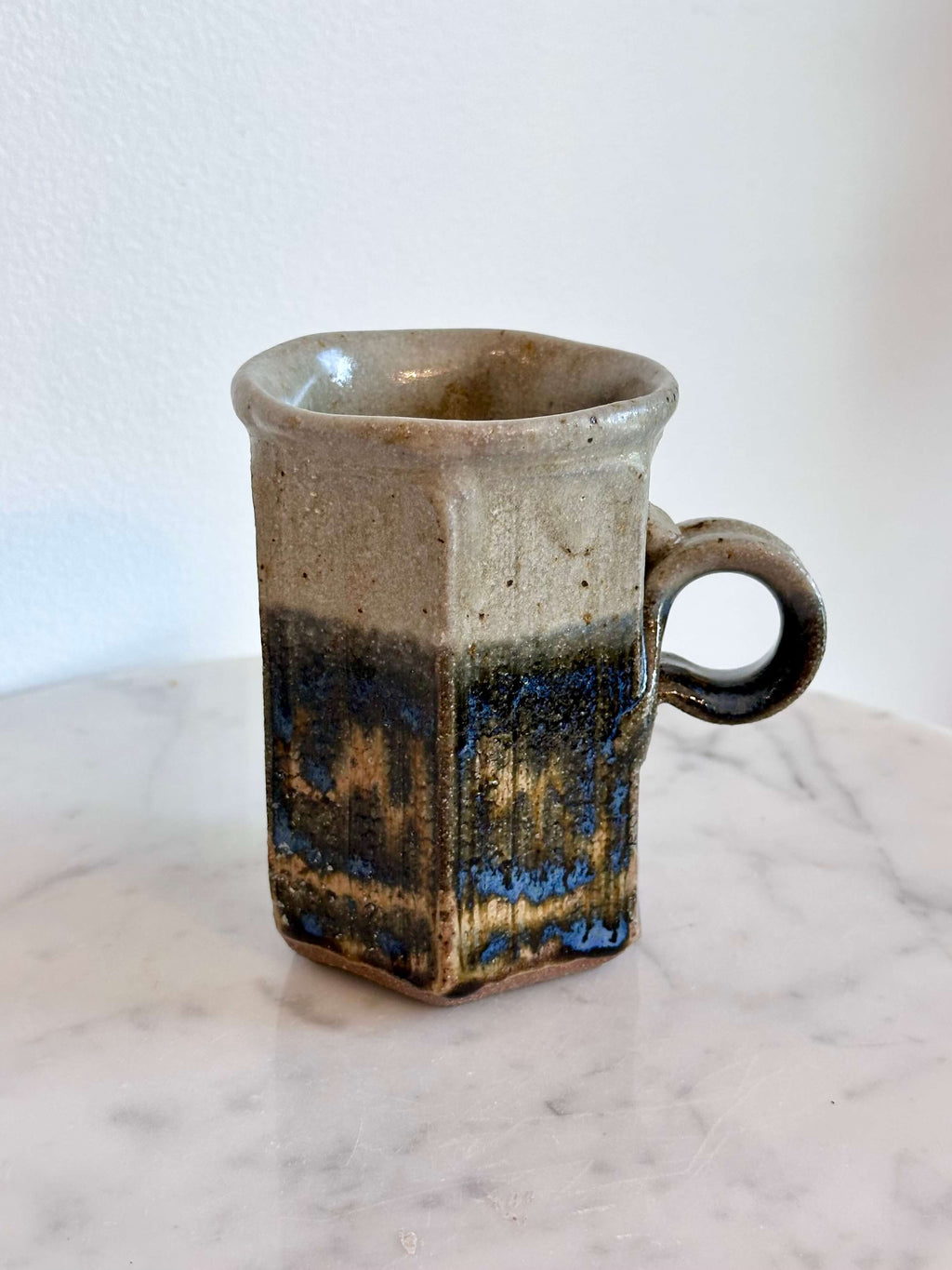 A handmade ceramic mug with a hexagonal body and a loop handle, featuring a layered glaze in gray, blue, and brown.