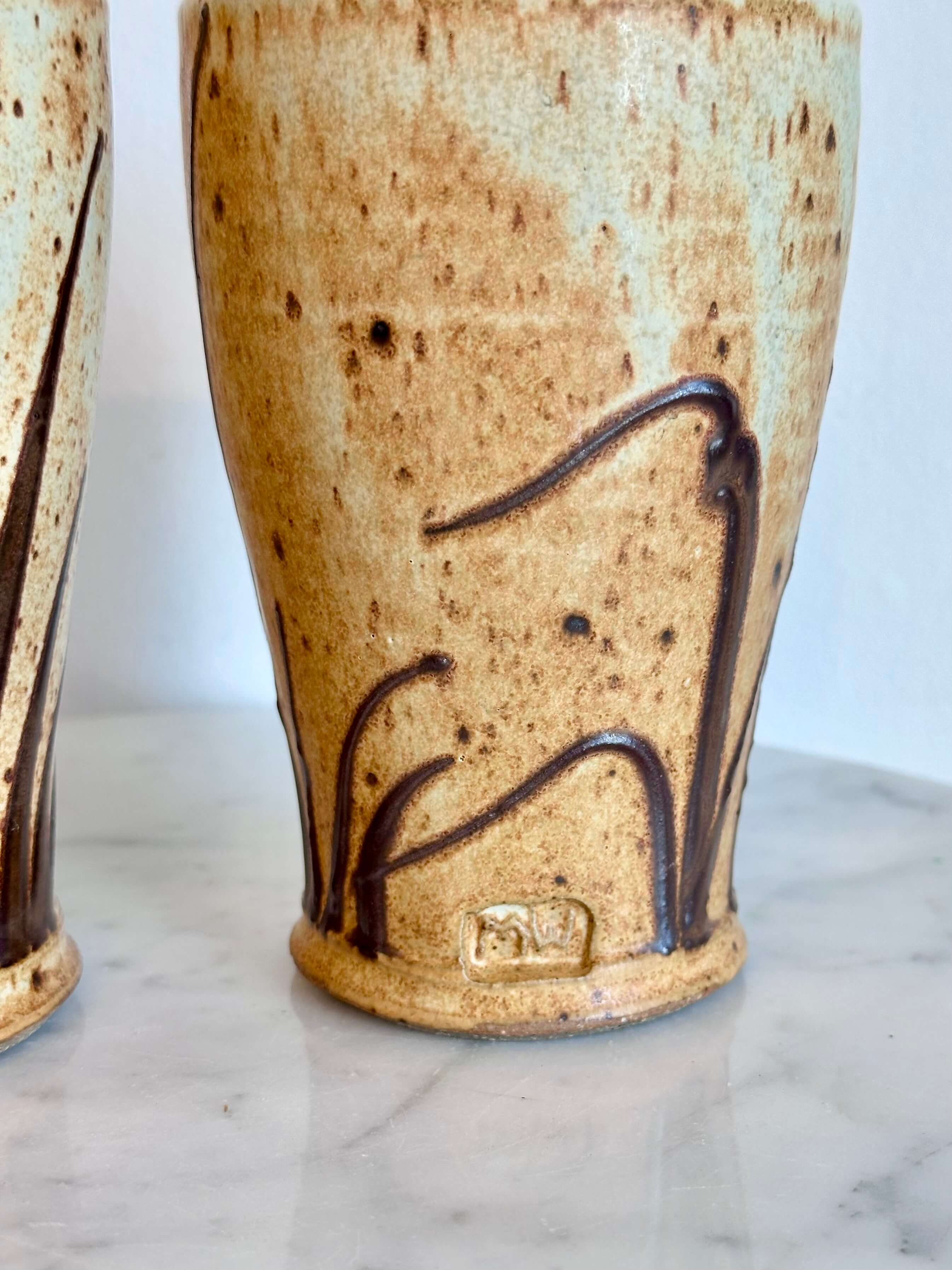 A close-up of a handmade ceramic vase with a textured beige and brown glaze, featuring dark brown abstract line details and a small stamped maker's mark near the base, set on a light marble surface.
