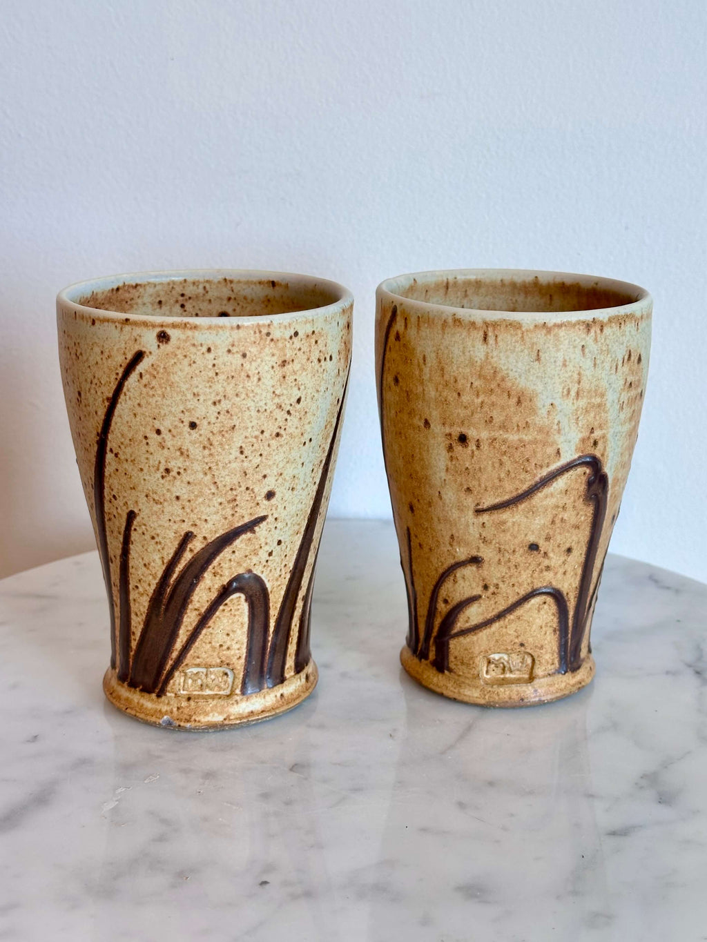 Two handmade ceramic cups with a speckled beige glaze and dark brown abstract grass-like patterns, displayed on a white marble surface against a white wall.