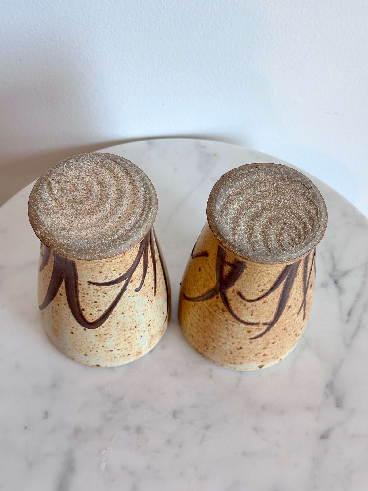 Two handmade ceramic cups with a speckled beige and brown glaze, decorated with dark brown abstract leaf-like patterns, shown on a white marble surface against a white wall.