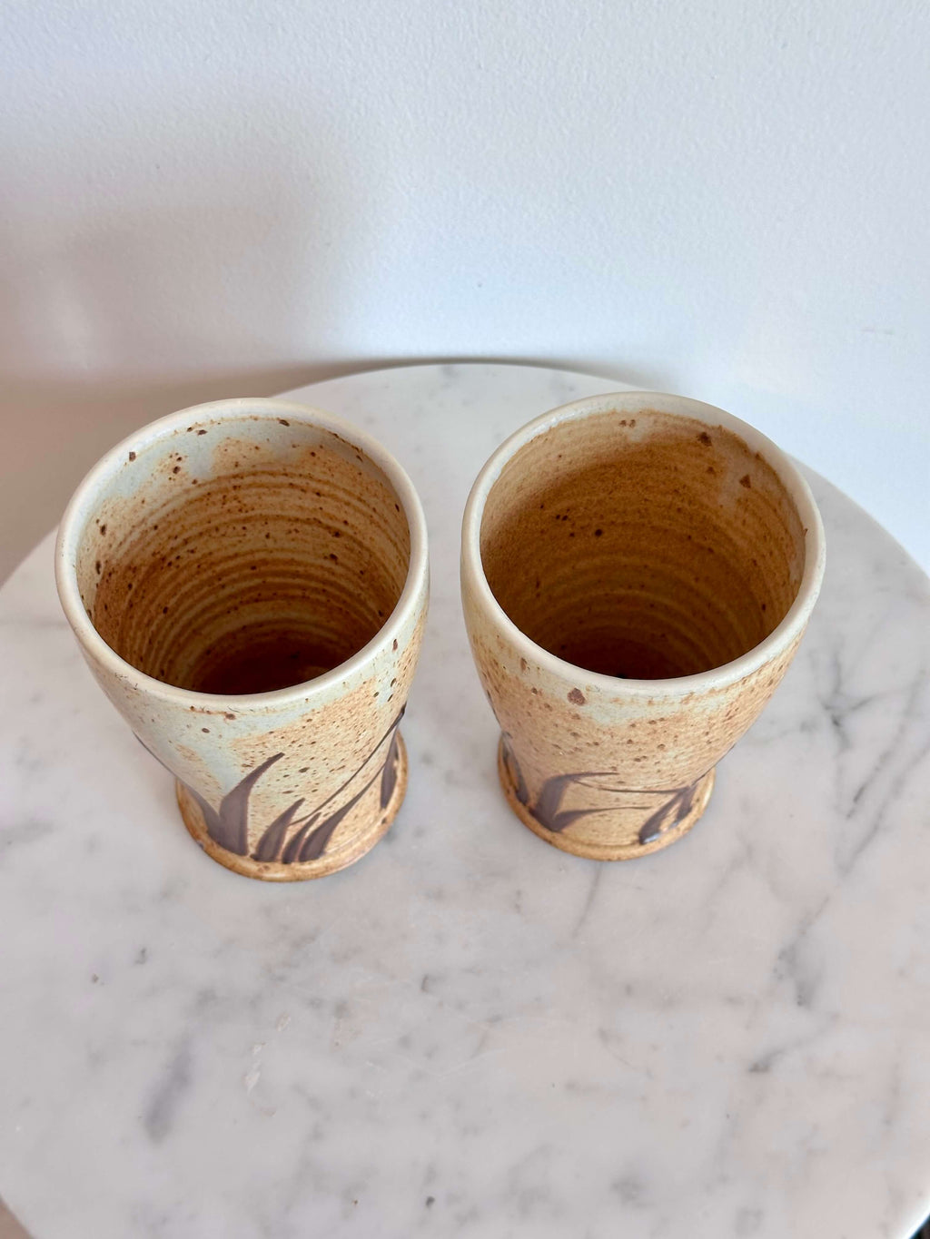Two handmade ceramic cups with a speckled beige and brown glaze, displayed side-by-side on a white marble surface against a white wall.