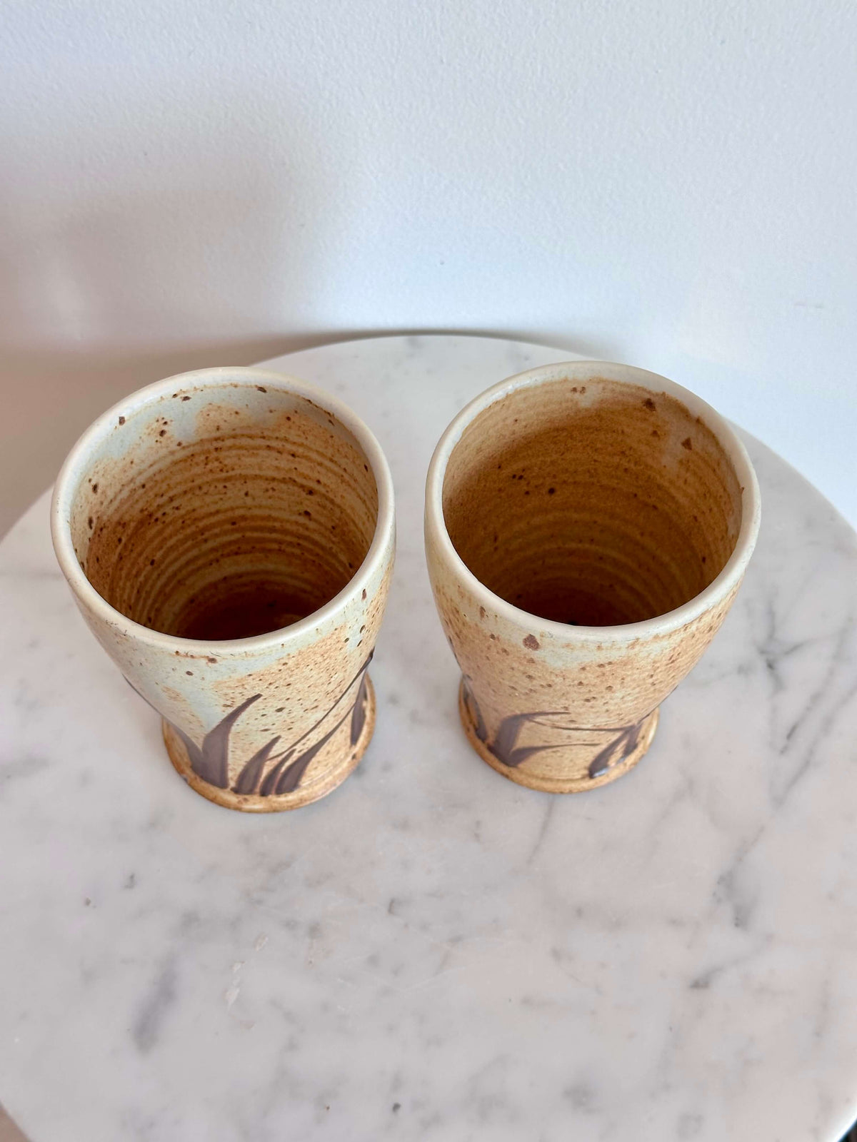 Two handmade ceramic cups with a speckled beige and brown glaze, displayed side-by-side on a white marble surface against a white wall.