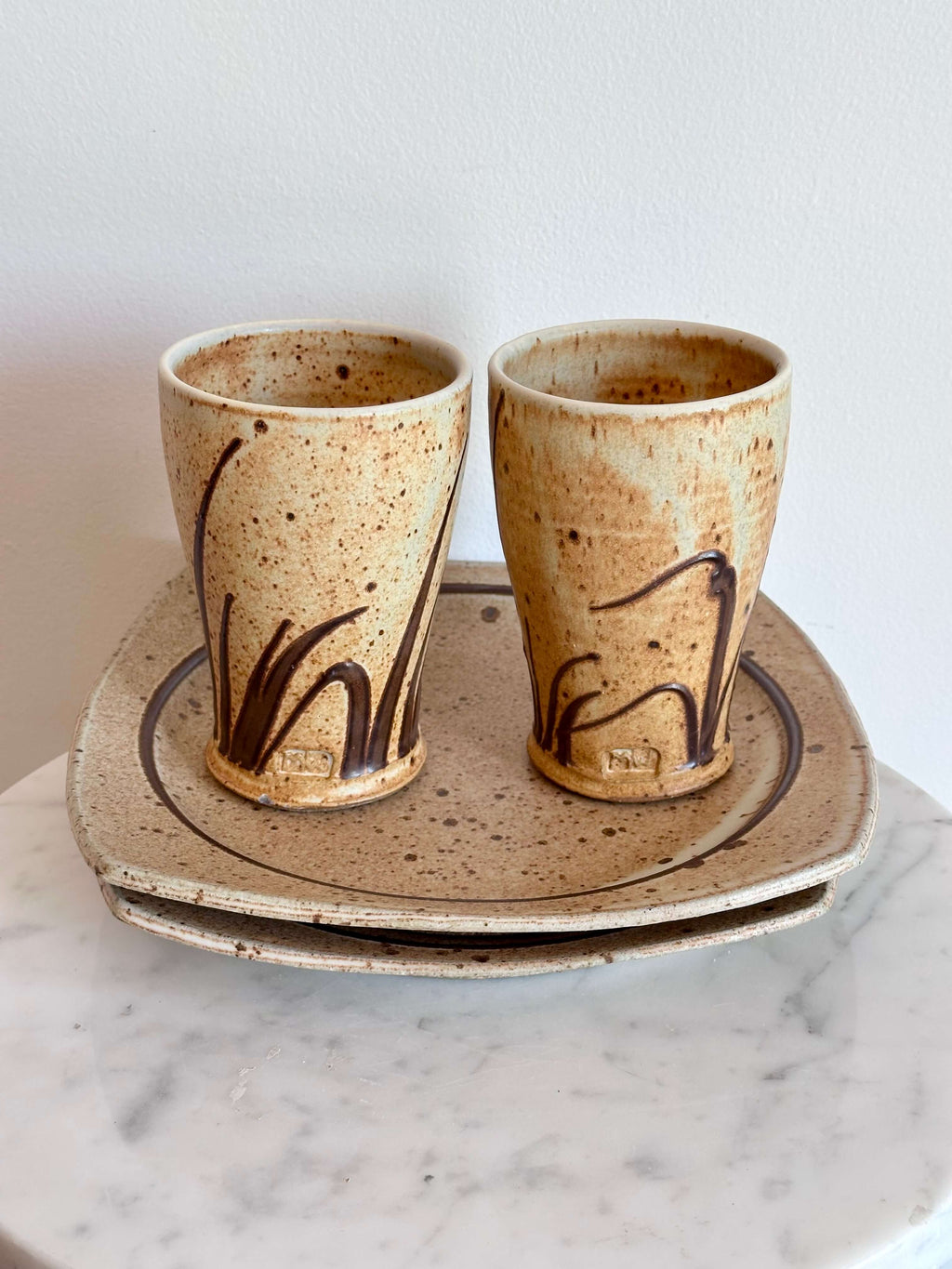 Two handmade ceramic cups with a speckled beige glaze and dark brown abstract brushstrokes, shown on a matching stack of ceramic plates against a white wall.