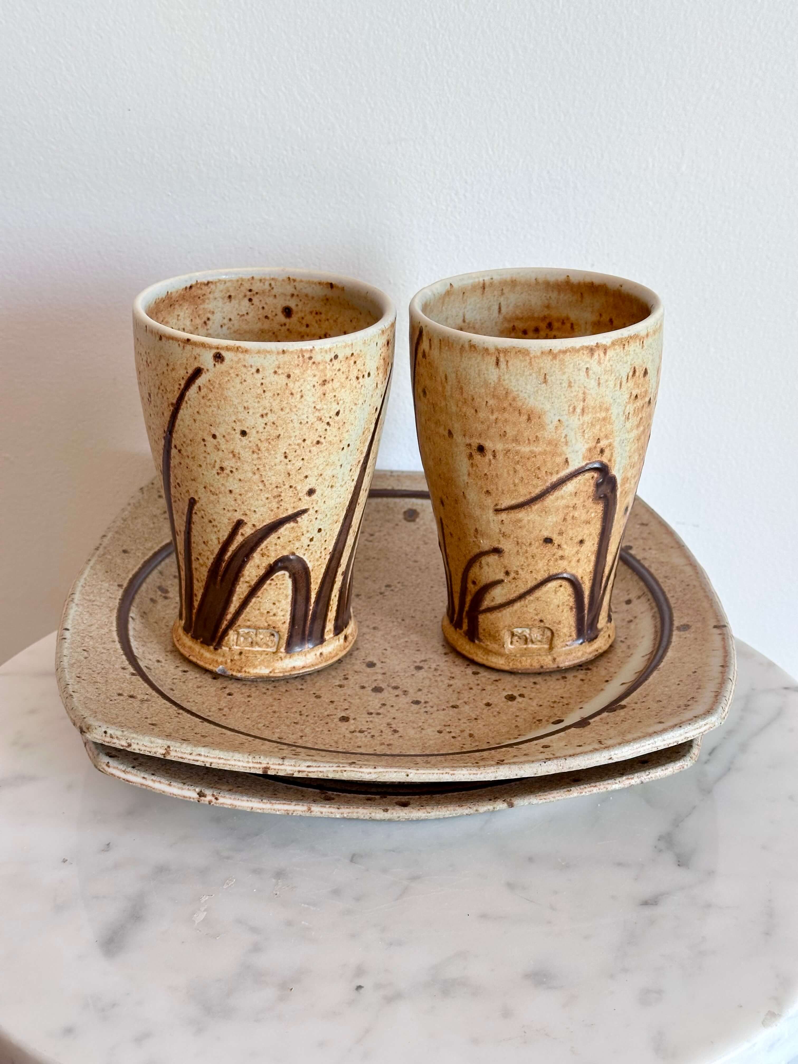 Two handmade ceramic cups with a speckled beige glaze and dark brown abstract brushstrokes, shown on a matching stack of ceramic plates against a white wall.