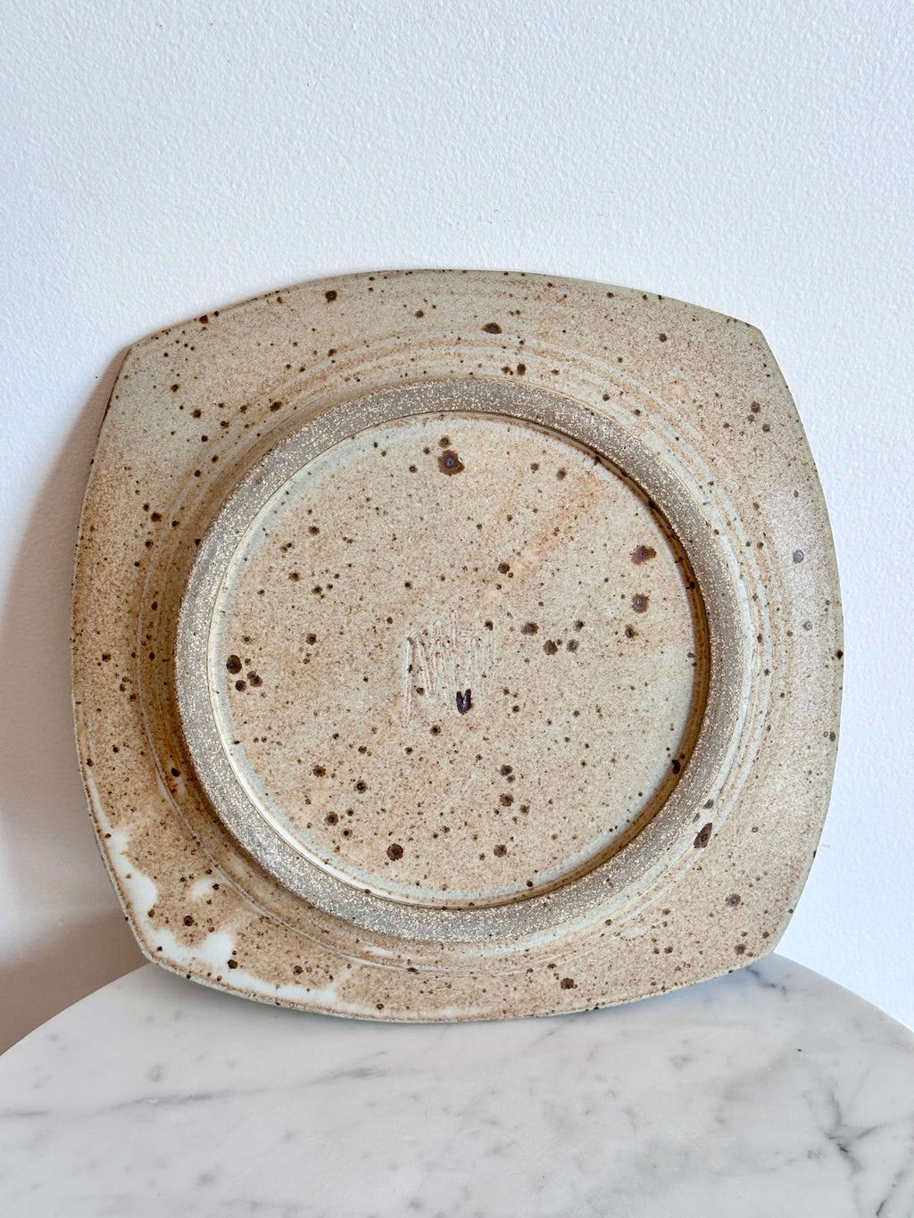 Back side of a square ceramic plate with rounded corners, featuring a speckled beige and brown glaze, displayed on a white marble surface against a white wall.