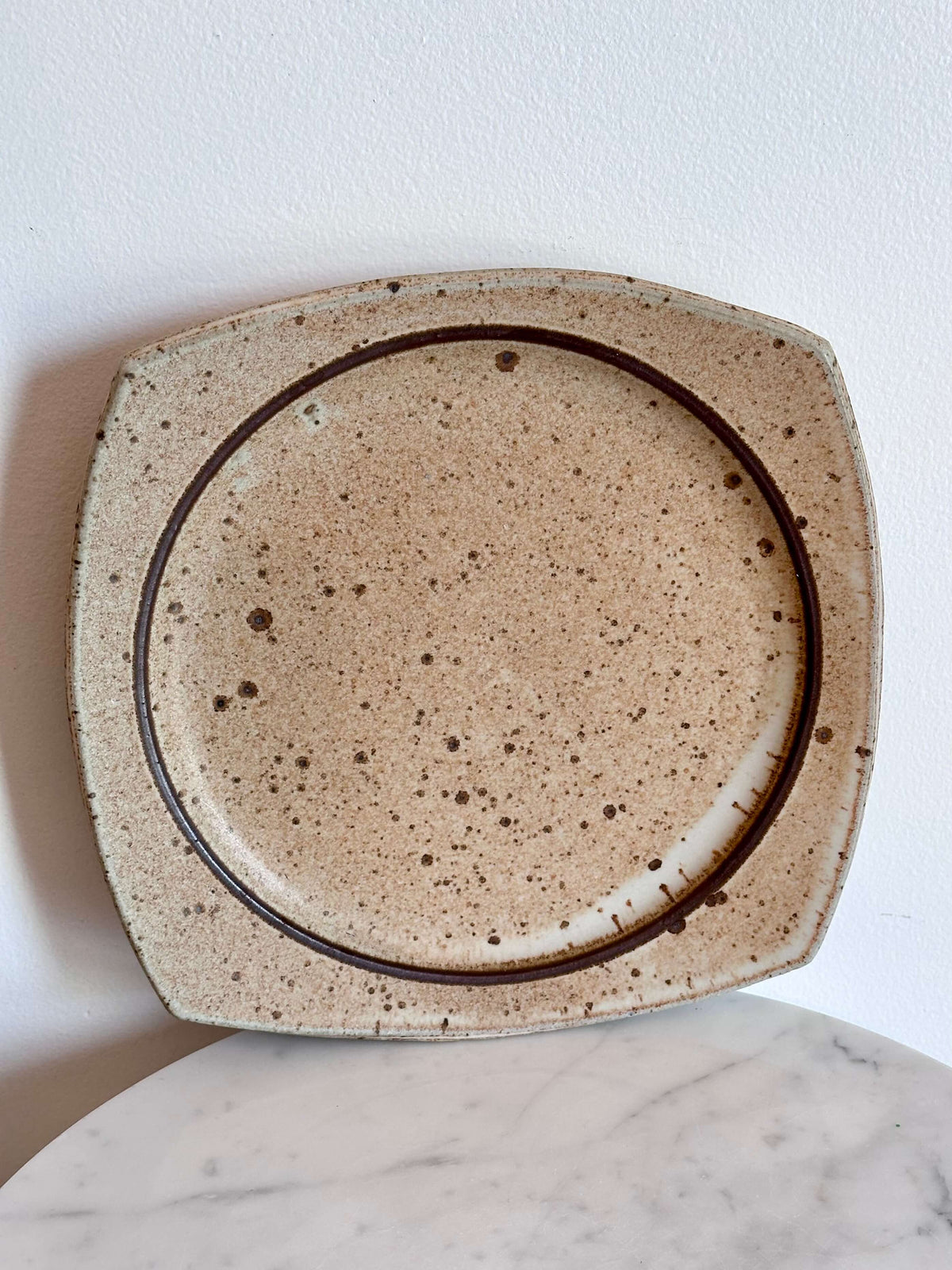 A square ceramic plate with rounded corners, featuring a speckled beige and brown glaze and a dark brown circular line design inside, shown against a white wall.