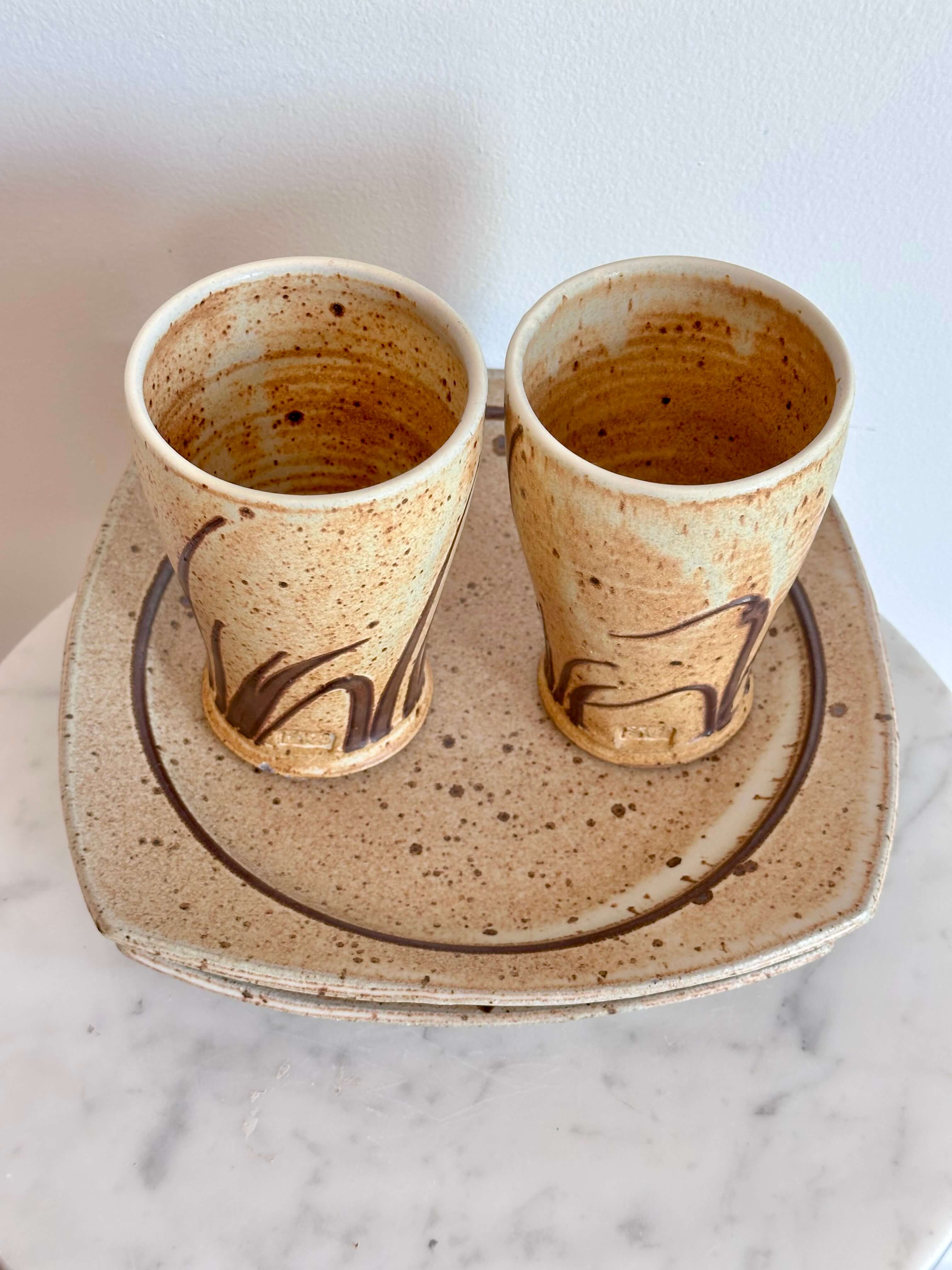 Two handmade ceramic cups with a speckled beige and brown glaze, decorated with dark brown abstract brushstrokes, sitting on a matching speckled ceramic plate.